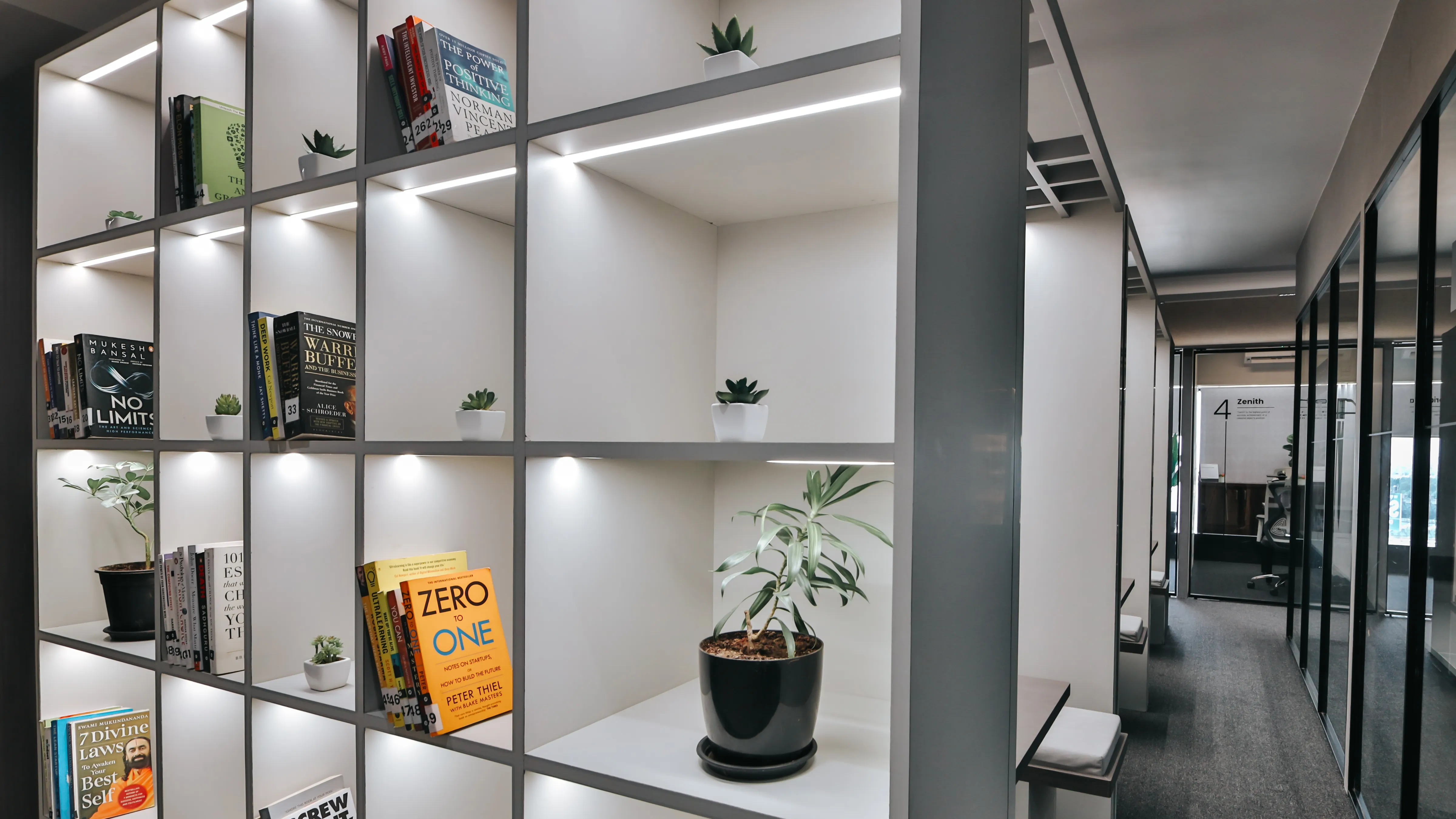 Modern shared office bookshelf with LED-lit cubicles, business books, and indoor plants in a premium coworking workspace