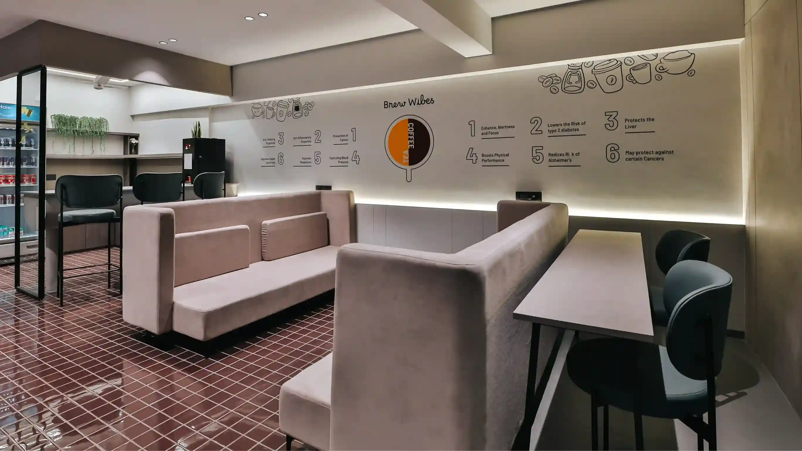 Modern office café or breakout lounge area with booth seating, a long counter table with chairs, warm ambient lighting, and a wall graphic explaining coffee benefits, creating a relaxed workspace environment.