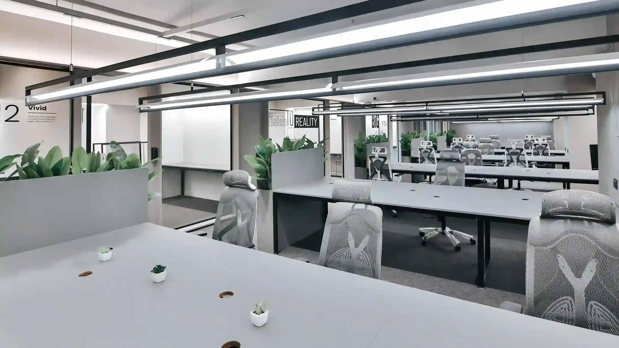 Spacious open coworking workspace with long shared desks, ergonomic office chairs, indoor plants, and modern lighting in a contemporary office environment.