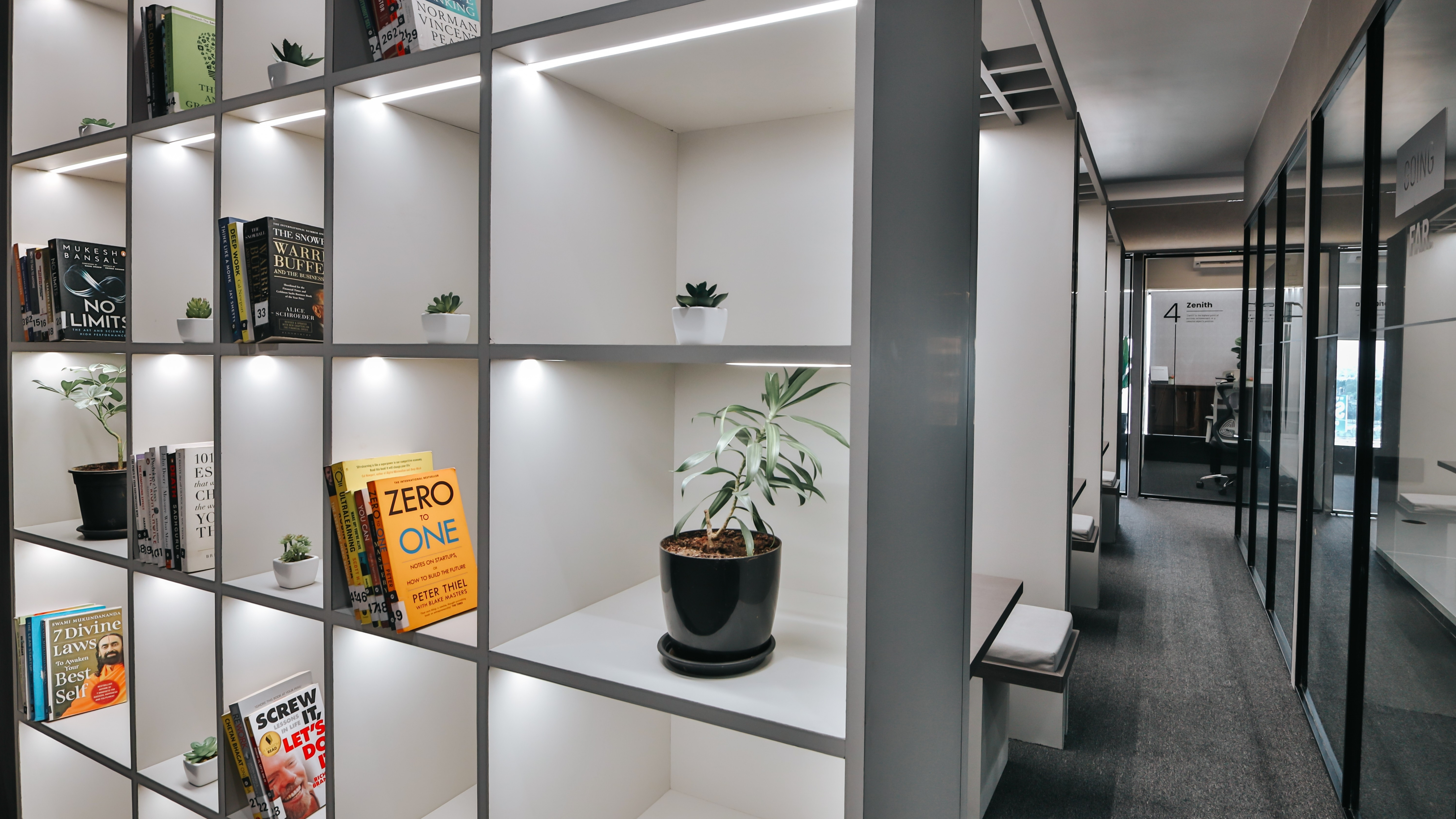 Modern office bookshelf with neatly arranged business books, indoor plants, and illuminated shelves in a minimalist coworking space.