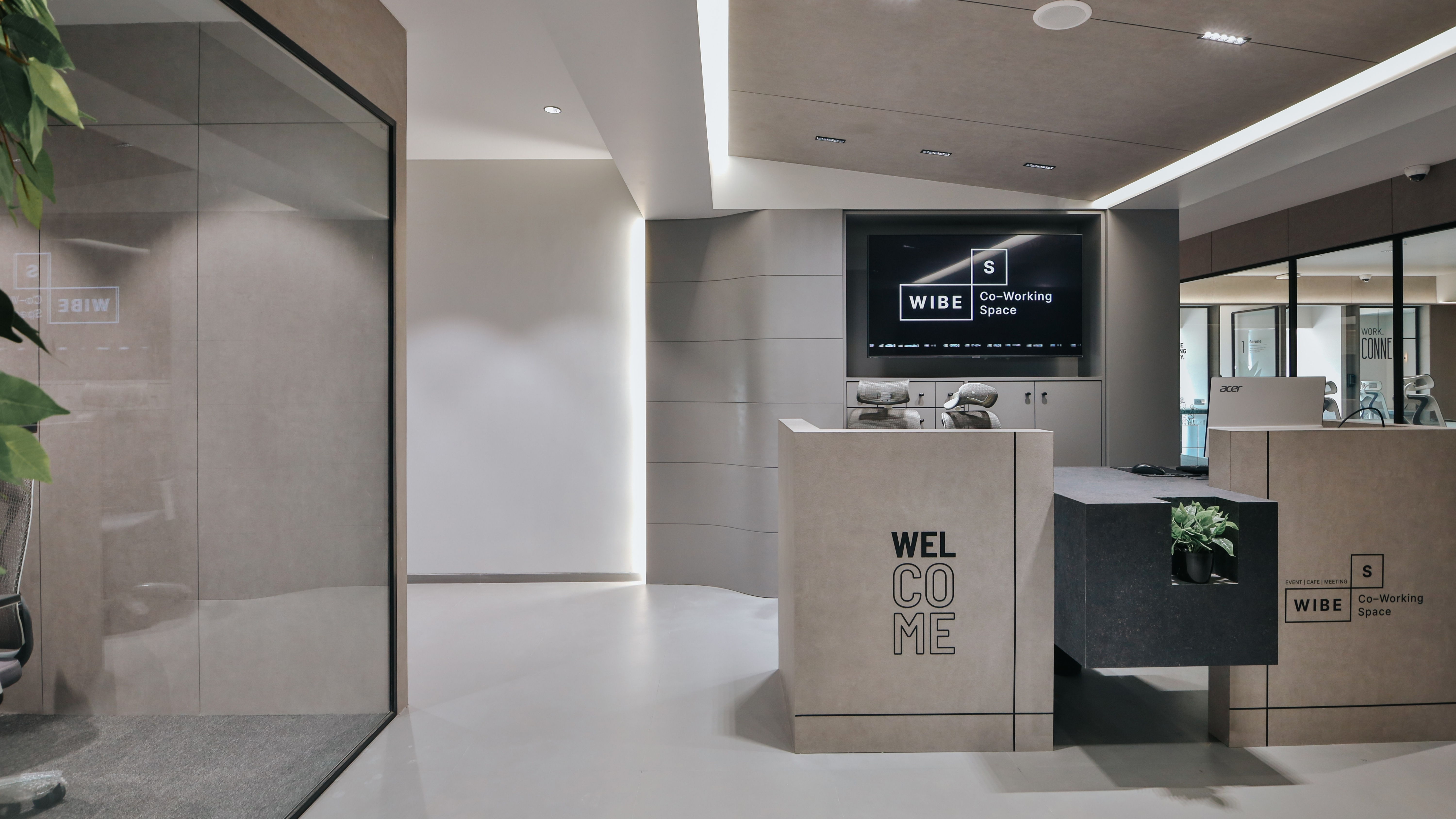Contemporary coworking office reception area with welcome desk, branding display screen, modern lighting, and minimal interior design.