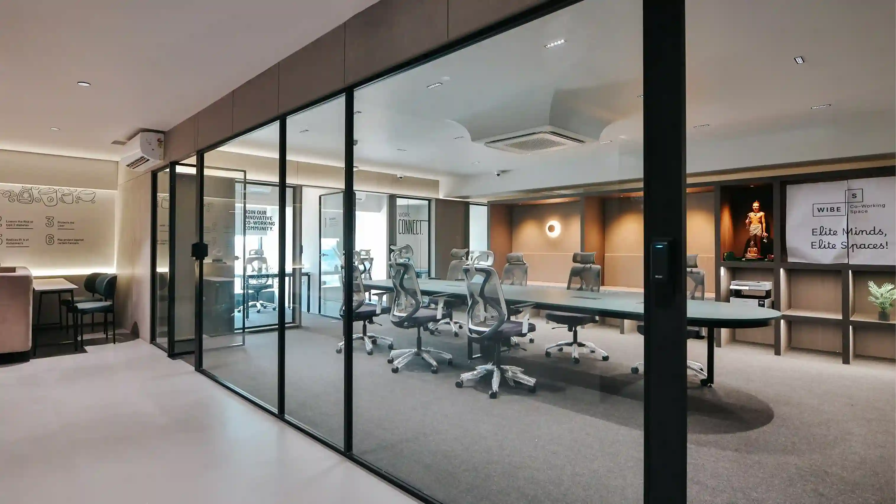 આધુનિક કો-વર્કિંગ સ્પેસમાં કાચની દીવાલવાળું મિટિંગ રૂમ, લાંબા કોન્ફરન્સ ટેબલની આસપાસ આરામદાયક ઑફિસ ખુરશીઓ, ગરમ લાઇટિંગ અને સ્વચ્છ, મિનિમલ ઇન્ટિરિયર ડિઝાઇન દર્શાવવામાં આવી છે.