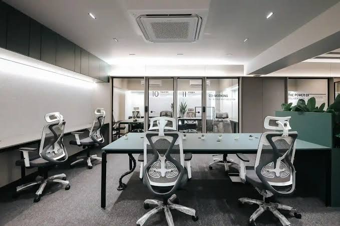 Modern coworking meeting room featuring a long teal conference table, ergonomic black-and-white office chairs, glass-partitioned meeting rooms, built-in wall storage, and bright contemporary lighting in a professional workspace.