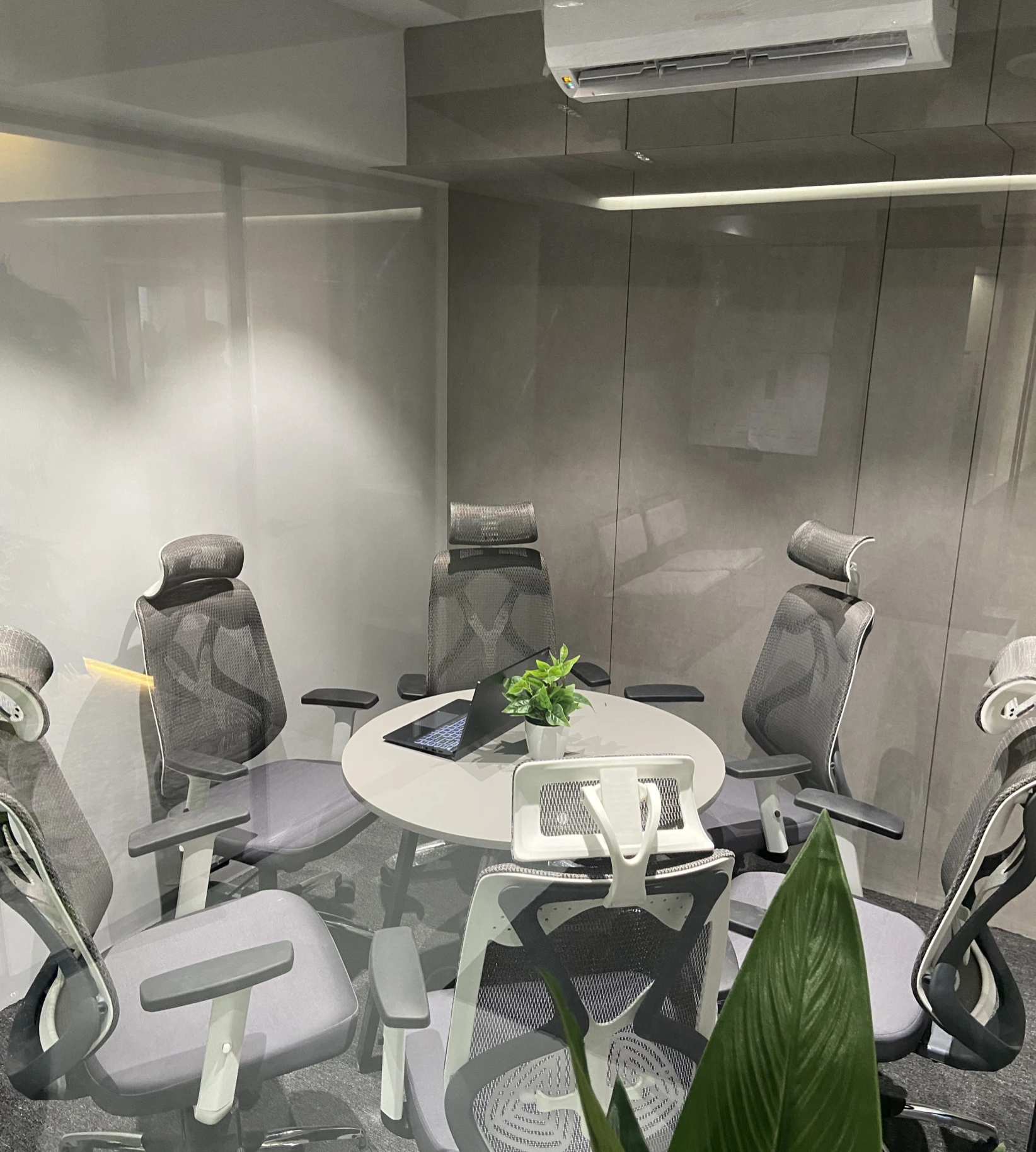 Contemporary office workspace featuring a large shared desk, ergonomic chairs, floor-to-ceiling windows, indoor plants, and a comfortable seating area.