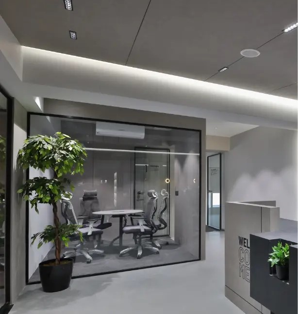 Modern coworking space reception with a glass-walled meeting room, ergonomic office chairs, indoor plants, and a minimalist welcome desk.