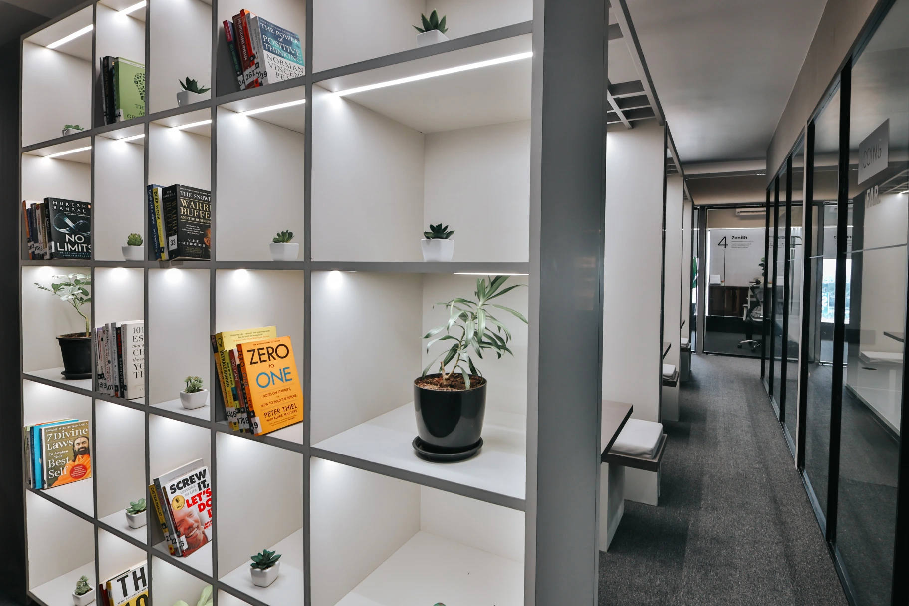 Modern coworking office corridor with illuminated bookshelves, indoor plants, glass meeting rooms, and contemporary workspace design.