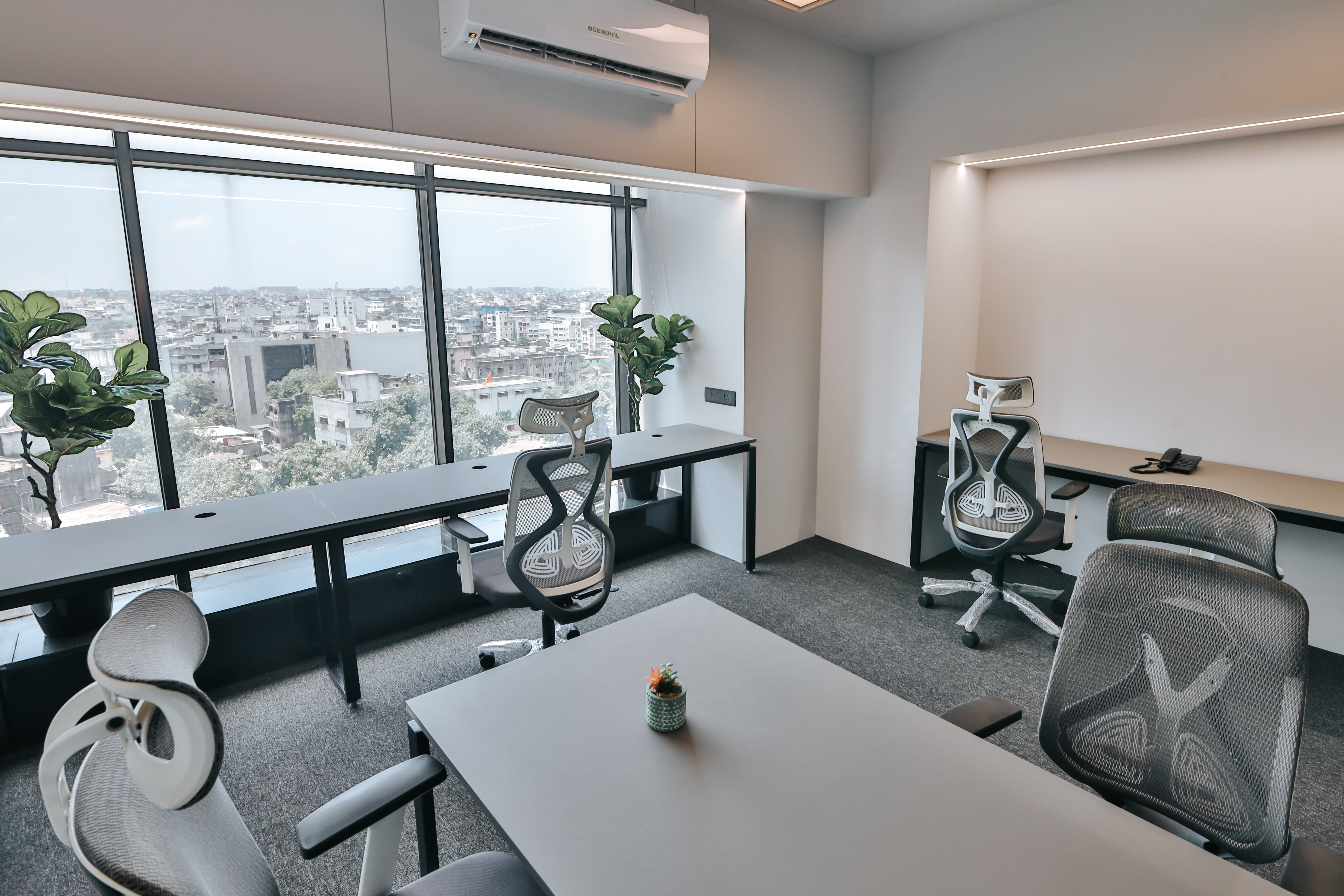 Modern office workspace with ergonomic chairs, long desks, large windows overlooking the city, indoor plants, and a wall-mounted air conditioner.