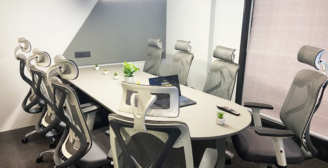 Modern meeting room with a long white conference table, ergonomic mesh chairs, a laptop, small potted plants, and large windows.